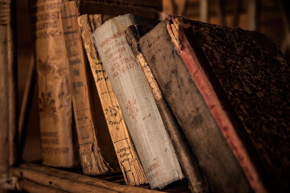 Stacks of old books, historical documents, and archived papers arranged inside a dimly lit research archive filled with history and memory.